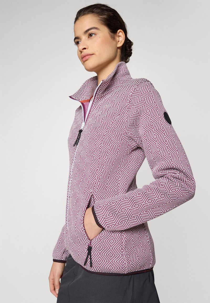 Giacca in pile con motivo geometrico rosa e bianco, colletto alto, chiusura frontale con zip e tasche laterali. Accenti neri ai polsini e sulle spalle.