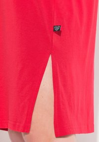 Robe en coton rouge avec fentes latérales, dotée d'un ourlet plié et d'une petite étiquette en tissu noir au bas de la couture. Texture lisse et design décontracté.