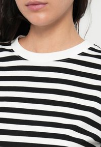 Melna un balta svītraina adīta blūze ar noapaļotu kakla izgriezumu un īsām piedurknēm, izceļas ar mīkstu tekstūru un vienmērīgu svītru rakstu.