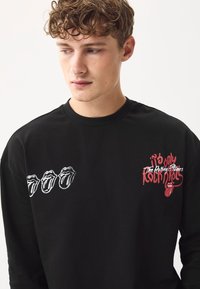 Sort langærmet t-shirt med hvide og røde Rolling Stones-grafikker. Blødt bomuldsstof med åben rund halsudskæring og afslappet pasform.