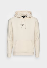 Beige hoodie i mjukt material med framficka, dragsko i huvan och en diskret svart logotyp på bröstet.