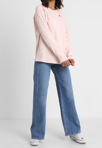 Lyserød langærmet T-shirt med rund hals, med et lille logo på brystet, parret med blå vidde denim jeans og hvide sneakers.