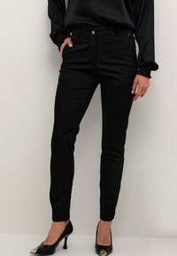 Pantalon noir slim à la texture lisse, doté de poches avant et d'une fermeture à bouton métallique, associé à des chaussures à talons hauts noires.