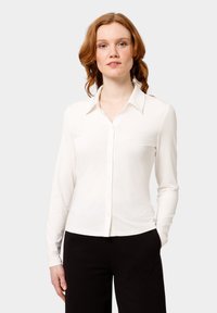 Camicia bianca a maniche lunghe con colletto, realizzata in tessuto morbido, caratterizzata da un design aderente e una leggera lucentezza. Abbinata a pantaloni neri.