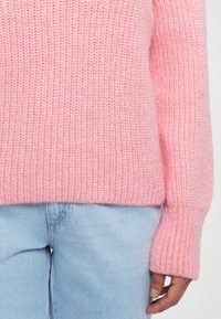 Osoba oblečená v ružovom hrubom pletenom svetri a svetlomodrých džínoch, ukazujúca strednú časť tela a pravú ruku uvoľnenú pri boku.