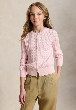 Ung person med langt blond hår som har på seg en pastellrosa kabelstrikket knappet cardigan og beige plisserte bukser, står med den ene hånden i lomma.