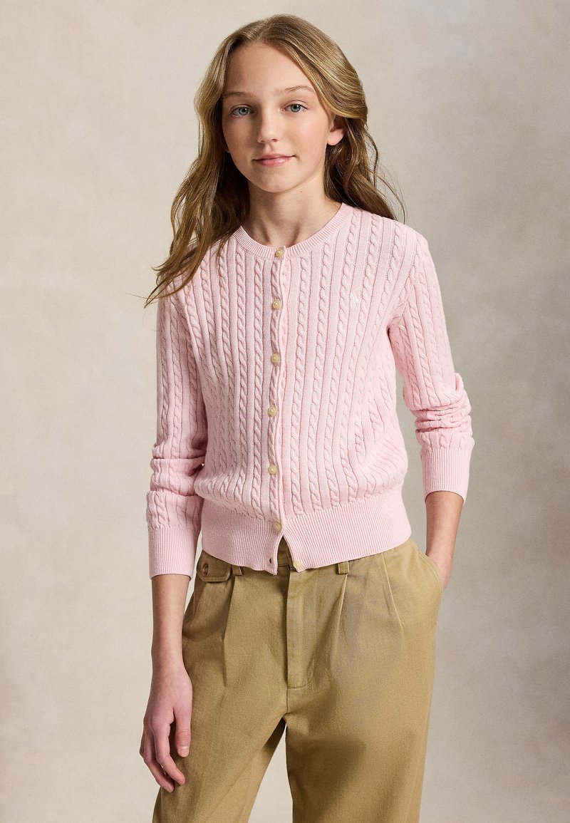 Hosszú szőke hajú fiatal személy, aki pasztell rózsaszín, fonott mintás, gombos kardigánt és bézs, pliszírozott nadrágot visel, egyik kezét a zsebében tartva áll.