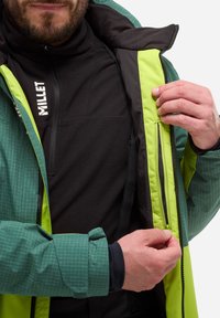Veste zippée verte et noire avec un motif en grille, dotée d'une doublure noire, de poignets ajustables et d'une poche sur le côté. Logo de la marque visible.