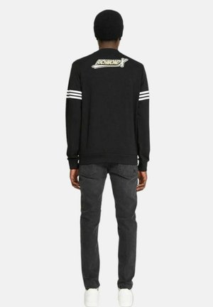 Felpa nera a maniche lunghe con strisce bianche sulle spalle, con il logo "RICHMOND X" sulla schiena. Abbinata a jeans scuri e scarpe bianche.