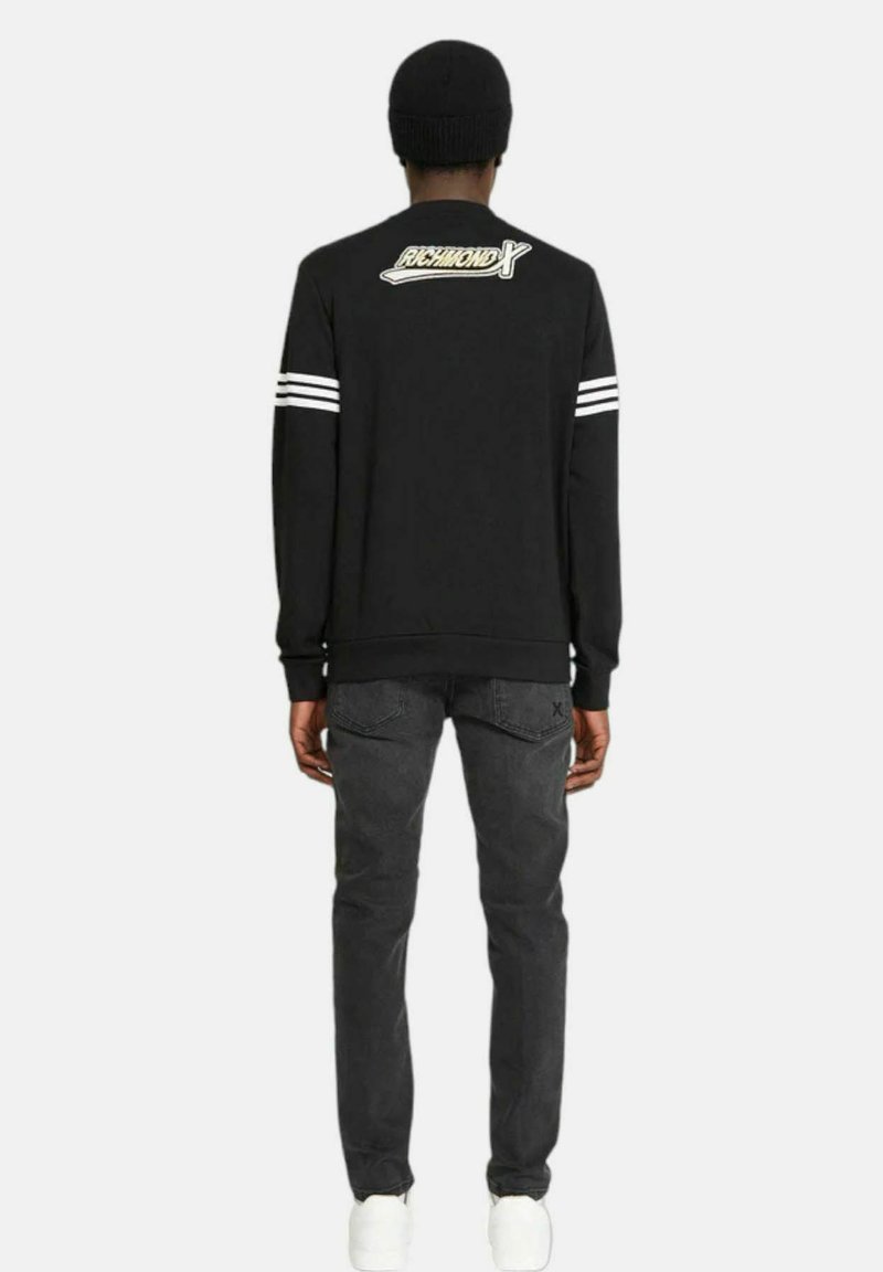 Felpa nera a maniche lunghe con strisce bianche sulle spalle, con il logo "RICHMOND X" sulla schiena. Abbinata a jeans scuri e scarpe bianche.