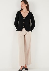 Black cardigan with a V-neckline, featuring a starburst brooch and golden buttons. Paired with beige wide-leg trousers and black heels.
