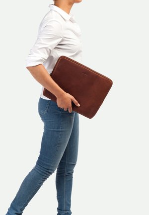 Funda para laptop de cuero marrón, forma rectangular, textura suave, cuenta con un zipper, sostenida con una mano. Complementada con una camisa blanca y unos jeans azules.