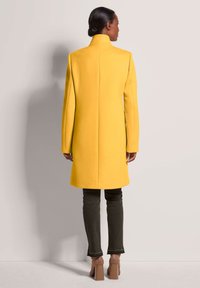 Geel wollen jas met hoge kraag, op maat gemaakt ontwerp en lange mouwen. Gepaard met donkergroene jeans en beige plateau schoenen.