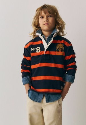 Blondt barn iført marineblå og orange stribet rugbytrøje over en denimskjorte, beige bukser, hænderne i lommerne, stående mod en ensfarvet hvid væg.