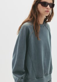 Izbljedjela teal dukserica s okruglim izrezom, rebrastim manžetama i bočnim šavovima. Mekana tekstura i opušteni kroj, u kombinaciji s tamnim naočalama.