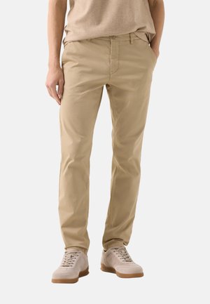 Pantalon beige à jambes droites porté avec des baskets beige et un haut beige assorti, les mains détendues sur les côtés et dans la poche.