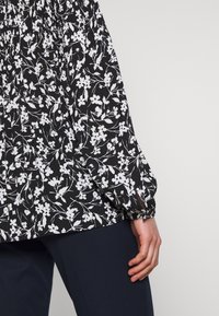 Black blouse with white floral print, featuring long, gathered sleeves with a cinched cuff and a loose fit. Worn over navy pants.