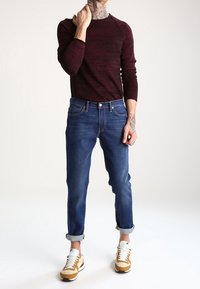 Camisa de manga larga a rayas en rojo oscuro combinada con unos vaqueros azules con los puños enrollados y zapatillas amarillas-marrones con acentos blancos. Antebrazo tatuado visible.