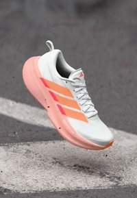 Λευκό αθλητικό παπούτσι με ελαφρύ σχεδιασμό. Διαθέτει ροζ λεπτομέρειες και τρεις λωρίδες στο πλάι. Υφή στη σόλα και διαπνέον υλικό.