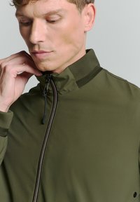 Olijfgroene zip-up jas met staande kraag, getextureerde accenten bij de hals en manchetten, en een gladde stof. Beschikt over een ritssluiting en subtiele details.