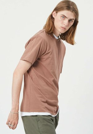 Brown short-sleeve shirt made of cotton, featuring a crew neckline and a relaxed fit. Layered over a white shirt. Green pants partially visible.