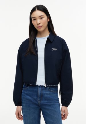 Młoda kobieta ubrana w granatową kurtkę zapinaną na zamek z logo "tommy jeans", jasnoniebieską koszulę i niebieskie dżinsy, stoi na białym tle.