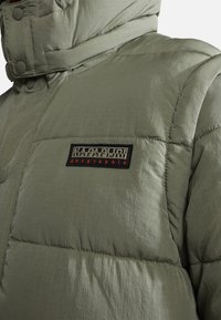 Olivengrüne Pufferjacke mit glatter, strukturierten Oberfläche. Enthält einen schwarzen Aufnäher mit dem "Napapijri"-Logo auf der Brust.