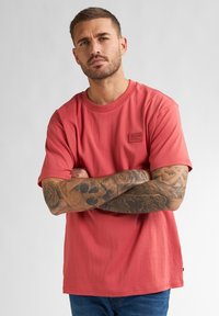 Koraalrood T-shirt met korte mouwen en een ronde halslijn, voorzien van een klein logopatch op de borst; model heeft getatoeëerde armen gekruist over zijn torso.