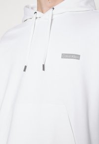 Felpa bianca con cappuccio regolabile, dotata di un'etichetta grigia con il logo Calvin Klein sul petto e una tasca frontale. Tessuto morbido al tatto.