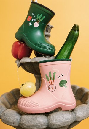 Børne-størrelse pink og grønne gummistøvler med grøntsagsillustrationer, der holder en zucchini og tomater, vist på en stenspringvand.
