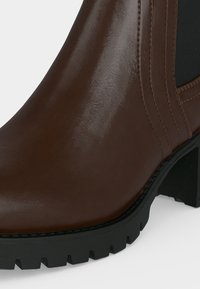 Gros plan d'une bottine en cuir marron avec un panneau latéral élastique noir, une semelle crantée épaisse et un talon bloc de hauteur moyenne.