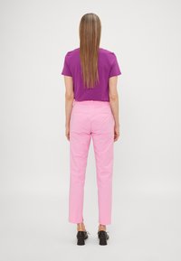Vrouw met lang, steil haar, gekleed in een paarse top met korte mouwen, roze broek en zwarte hakken, staande met haar rug naar de kijker op een effen achtergrond.