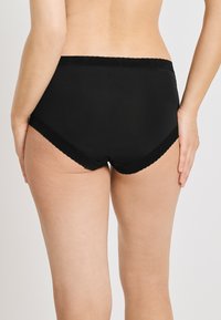 Bakvyn av en person som bär svart högmidjad underkläder med spetskant vid midjan och benöppningarna mot en neutral bakgrund.