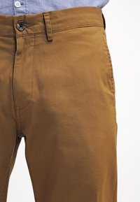 Braune Baumwollhose mit glatter Textur, ausgestattet mit Reißverschluss, Knopfverschluss und vorderen Taschen. Lockerer Schnitt mit einfachem Design.