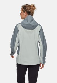 Lichtgrijze waterdichte jas met een capuchon, donkerder grijze accenten op de schouders en de mouwen, gestructureerde stof en een logo op de achterkant.