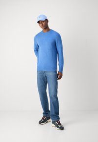 Blauer Strickpullover mit Rundhalsausschnitt, kombiniert mit einer hellblauen Mütze, blauen Jeans und mehrfarbigen Sneakers. Weiche Textur, lässiger Schnitt.