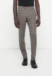 Szürke kockás nadrág, testre szabott szabással, diszkrét narancssárga akcentussal. Sima tapintású anyagból készült. Fekete cipők egészítik ki a megjelenést.