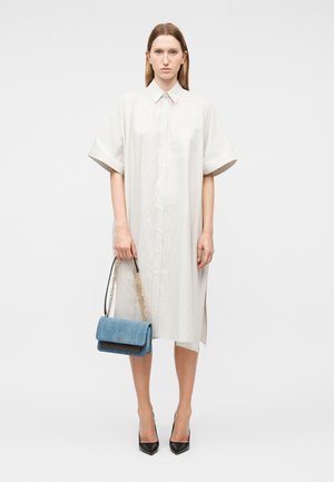 Γυναίκα με χαλαρό λευκό φόρεμα που φτάνει μέχρι το γόνατο, κρατώντας μπλε τσάντα με χρυσή αλυσίδα, φορούντας μαύρες μυτερές γόβες, στέκεται σε λευκό φόντο.