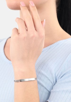 Mujer con un top de canalé azul claro tocándose la barbilla, lleva una pulsera de cadena de plata con una placa rectangular en blanco en su muñeca.