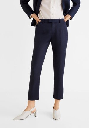 Femme portant un pantalon bleu marine ajusté à la cheville, une chemise blanche à col, un blazer bleu marine et des chaussures blanches à talons avec bride arrière, debout les mains dans les poches.