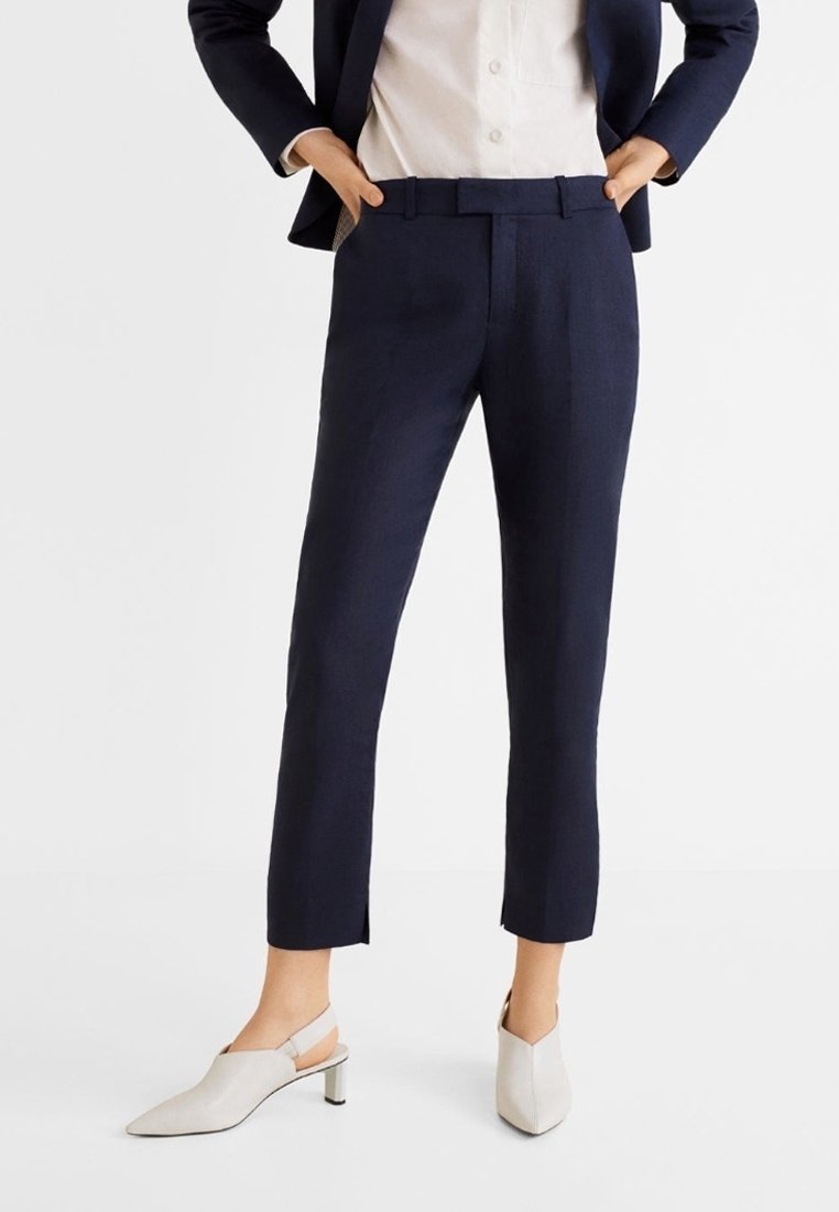 Femme portant un pantalon bleu marine ajusté à la cheville, une chemise blanche à col, un blazer bleu marine et des chaussures blanches à talons avec bride arrière, debout les mains dans les poches.