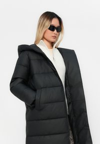 Sort pufferjakke med en stor hætte, lynlåsfront og sidelommer; har en quiltet struktur og en høj krave, båret over en cremet sweater.