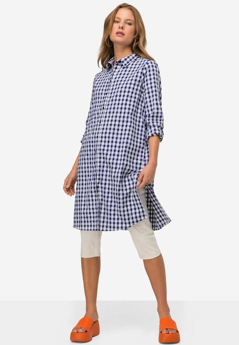 Abito camicia in gingham blu e bianco con bottoni sul davanti, maniche lunghe e orlo asimmetrico. Abbinato a leggings bianchi e zoccoli arancioni brillanti.