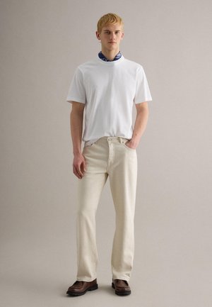 Jonge man die een wit T-shirt, beige broek, bruine laarzen en een blauwe halsaccessoire met patroon draagt, staand tegen een effen grijze achtergrond.