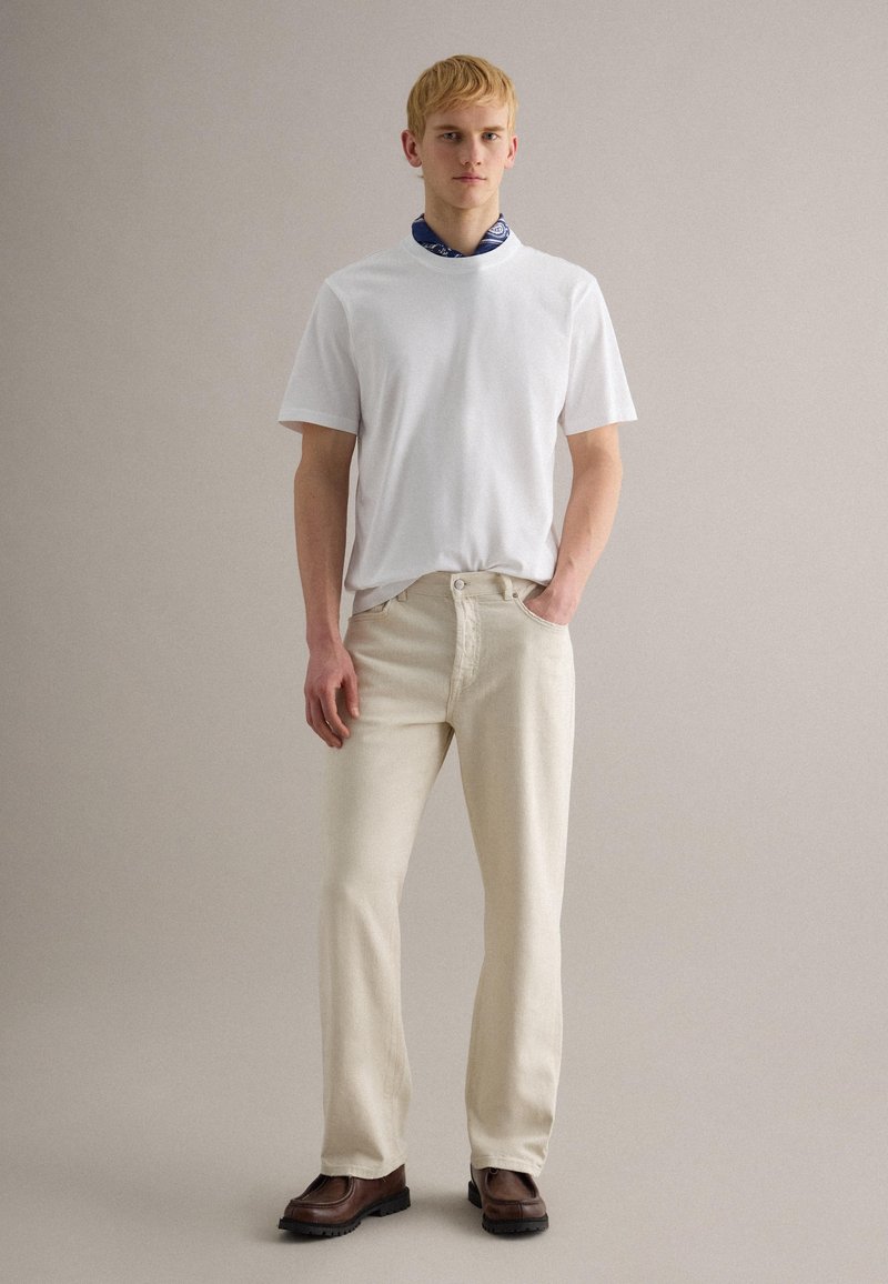 Giovane uomo che indossa una maglietta bianca, pantaloni beige, stivali marroni e una sciarpa al collo blu con motivi, in piedi davanti a uno sfondo grigio uniforme.