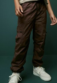 Braune Cargo-Hosen mit elastischen Bündchen, mehreren Taschen und einem Kordelzugbund, kombiniert mit weißen Nike Sneakern. Glatte Stoffoberfläche.