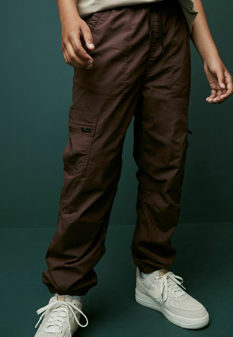 Braune Cargo-Hosen mit elastischen Bündchen, mehreren Taschen und einem Kordelzugbund, kombiniert mit weißen Nike Sneakern. Glatte Stoffoberfläche.
