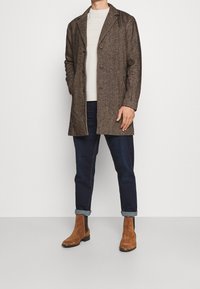 Brauner Fischgrätenmantel mit Reverskragen, kombiniert mit einem hellen Pullover, dunklen hochgekrempelten Jeans und braunen Wildleder-Stiefeletten.