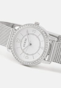 Montre avec un bracelet en mailles argentées, un cadran rond blanc, une lunette avec des accents en cristal et des index des heures argentés. Marque "Guess" affichée sur le cadran.