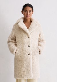 Beige, fluffy jas van een textuur die lijkt op schapenvacht, grote kraag en vijf zwarte knopen aan de voorkant; heeft zijzakken.